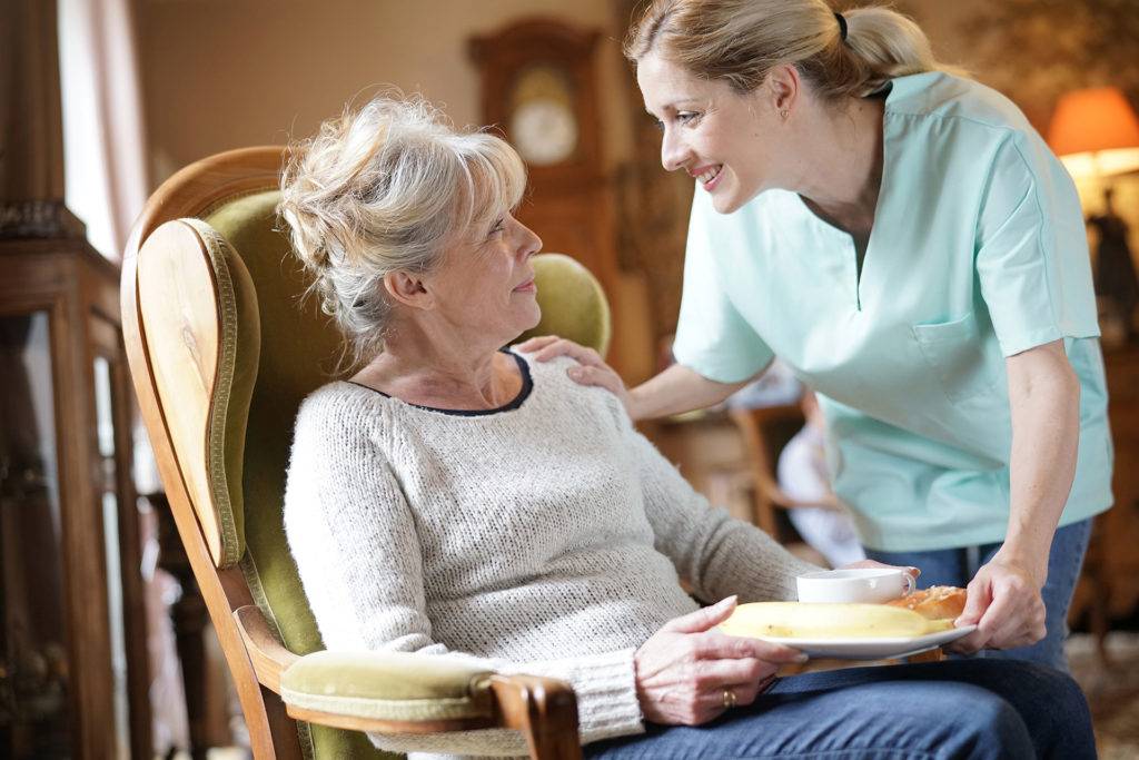 Caretaker Handing Breakfast to Senior Woman Sitting in Rocking Chair